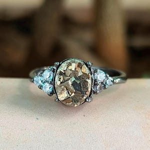 May include: A silver ring featuring an oval-shaped, textured, golden-brown gemstone as the centerpiece. The ring is flanked by clusters of small, clear gemstones on each side. The setting is dark, and the ring is displayed against a blurred background.