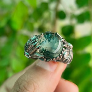 Può includere: Un anello d'argento con una grande pietra verde rotonda e traslucida con inclusioni verde scuro. L'anello ha un'incastonatura ornata e dettagliata con disegni a forma di foglia e piccole pietre chiare. L'anello è tenuto contro uno sfondo verde sfocato.