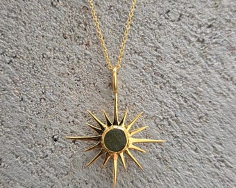 May include: A gold-colored necklace featuring a sunburst pendant with a central, circular, dark green stone. The pendant has multiple pointed rays extending outwards. The necklace chain is also gold-colored, and the background is a textured gray surface.