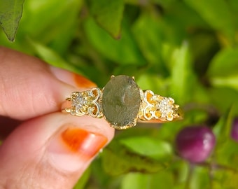 May include: A gold ring featuring an oval, olive-green gemstone. The ring has ornate filigree detailing on the band and small, clear accent stones on either side of the main stone. The ring is held against a backdrop of green foliage.