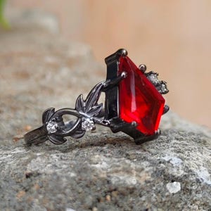 May include: A black metal ring featuring a kite-shaped red gemstone as the centerpiece. The band is designed with intricate leaf-like metalwork and is accented with small, clear, round-cut stones embedded along the sides of the setting.