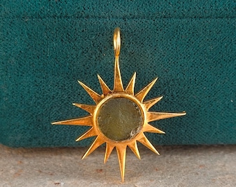 May include: A gold-colored pendant in the shape of a sunburst. The pendant features a central, circular, olive-green stone. The sunburst design has pointed rays extending outwards from the center. The pendant has a small loop at the top for a chain.