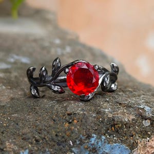 Op de afbeelding: Een ring met een donkere, gunmetal-kleurige band in de vorm van een wijnstok met kleine blaadjes. Een enkele, ronde, gefacetteerde, felrode edelsteen is in het midden van de band gezet en wordt vastgehouden door de metalen wijnstokstructuur.
