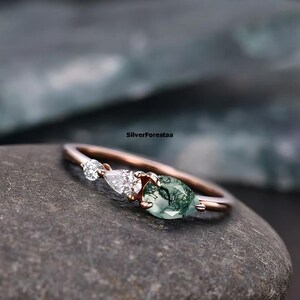 May include: A delicate rose gold ring with a green moss agate gemstone and two smaller clear pear-shaped gemstones. The ring is set on a gray stone background.