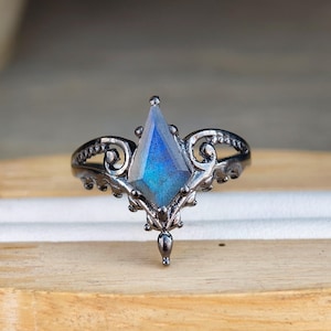Könnte beinhalten: Ein dunkel versilberter Ring mit einem großen, blauen Edelstein in Diamantenform. Der Ring hat ein verziertes, filigranes Design um den Edelstein und entlang des Rings, was ihm einen Vintage- oder Gothic-Look verleiht. Der Edelstein hat eine schimmernde, irisierende Qualität.