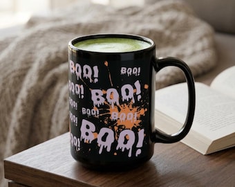 May include: Black ceramic mug with the word "BOO!" in white and purple, with an orange splatter design. The mug is filled with a green liquid. The mug is on a wooden surface.
