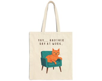 May include: Off-white canvas tote bag featuring a grumpy orange cat lounging in a teal armchair. The text "YAY... ANOTHER DAY AT WORK" is printed above the cat. The bag has two handles for carrying.