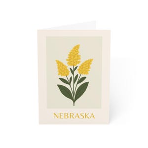 May include: Greeting card featuring a botanical illustration of yellow flowers with green leaves against a beige background. The word "NEBRASKA" is printed in gold at the bottom of the card.