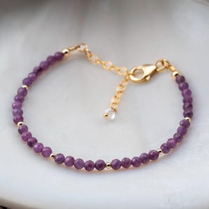 May include: A delicate bracelet featuring small, faceted purple beads interspersed with gold-colored beads. The bracelet has a gold-colored clasp and adjustable chain. The jewelry is displayed on a white surface.