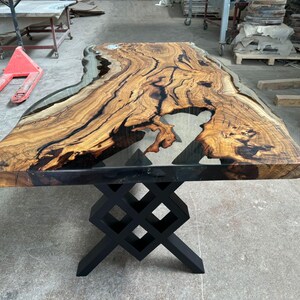 May include: A large wooden table with a live edge top and a clear epoxy resin fill. The table has a black metal base with a geometric design.