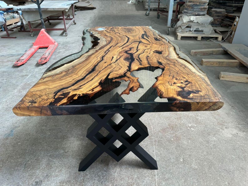 May include: A large wooden table with a live edge top and a clear epoxy resin fill. The table has a black metal base with a geometric design.