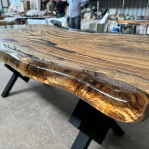 May include: Close-up of a live edge wood table top with a clear epoxy resin finish. The table top has a natural wood grain pattern and a dark brown color. The epoxy resin is clear and glossy, and it highlights the natural beauty of the wood.