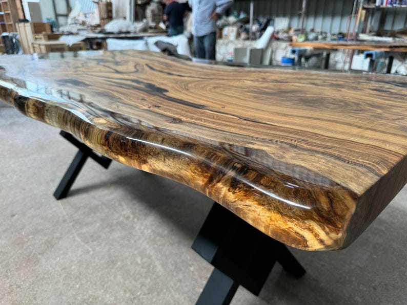 May include: Close-up of a live edge wood table top with a clear epoxy resin finish. The table top has a natural wood grain pattern and a dark brown color. The epoxy resin is clear and glossy, and it highlights the natural beauty of the wood.