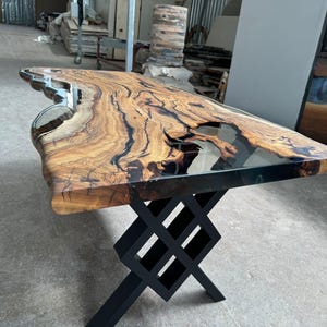 May include: A large wooden table with a live edge top and a clear epoxy resin fill. The table has a black metal base with a criss-cross design.