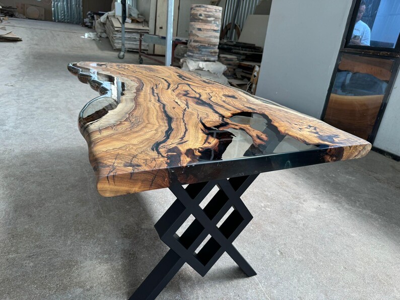 May include: A large wooden table with a live edge top and a clear epoxy resin fill. The table has a black metal base with a criss-cross design.