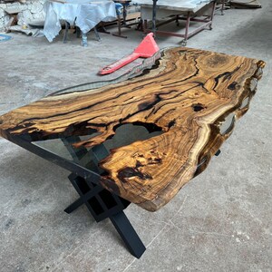 May include: A large wooden table top with a live edge and black epoxy resin filling in the gaps. The table top is supported by a black metal X-shaped base.