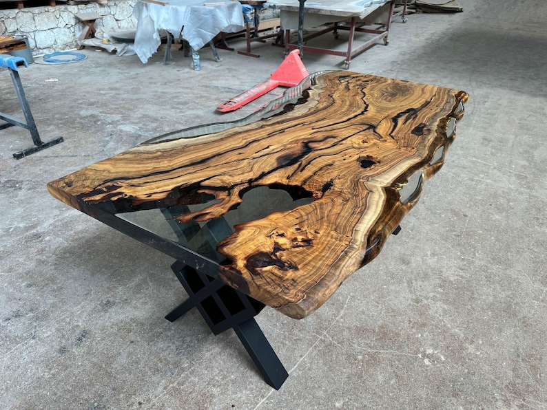 May include: A large wooden table top with a live edge and black epoxy resin filling in the gaps. The table top is supported by a black metal X-shaped base.