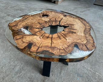 Könnte beinhalten: Ovaler Couchtisch mit einzigartigem Design. Die Tischplatte zeigt einen Querschnitt eines Baumstamms, der in klarem Harz eingebettet ist. Das Holz hat eine satte braune Farbe mit sichtbarer Maserung und einer großen zentralen Öffnung. Er steht auf schwarzen Metallbeinen. Ca. 120 cm lang.