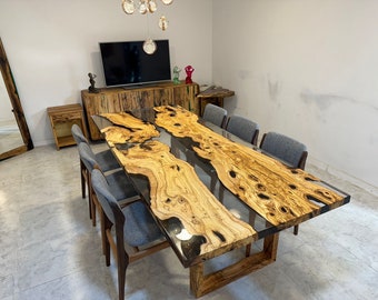 Op de afbeelding: Een grote houten eettafel met een inleg van transparante epoxyhars. De tafel heeft een natuurlijke houtkleur en is omringd door grijze stoelen.