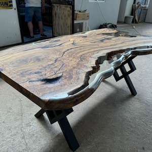 May include: A large wooden table with a live edge and a clear epoxy resin fill. The table has a black metal X-shaped base.