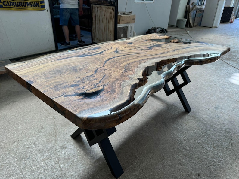 May include: A large wooden table with a live edge and a clear epoxy resin fill. The table has a black metal X-shaped base.