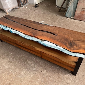Op de afbeelding: Een lange, houten tv-standaard met een live edge-blad en een blauwe harsinleg. De standaard heeft twee laden en een plank. De standaard is gemaakt van donker hout en heeft een natuurlijke, rustieke uitstraling.