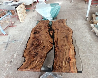Può includere: Un grande piano da tavolo in legno a bordo vivo con un riempimento in resina epossidica trasparente. Il legno è di colore marrone scuro con motivi di venatura naturali. La resina epossidica è trasparente e ha una finitura liscia.