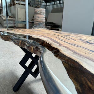 May include: A wooden table top with a live edge and a clear epoxy resin finish. The table top is supported by a black metal X-shaped base.
