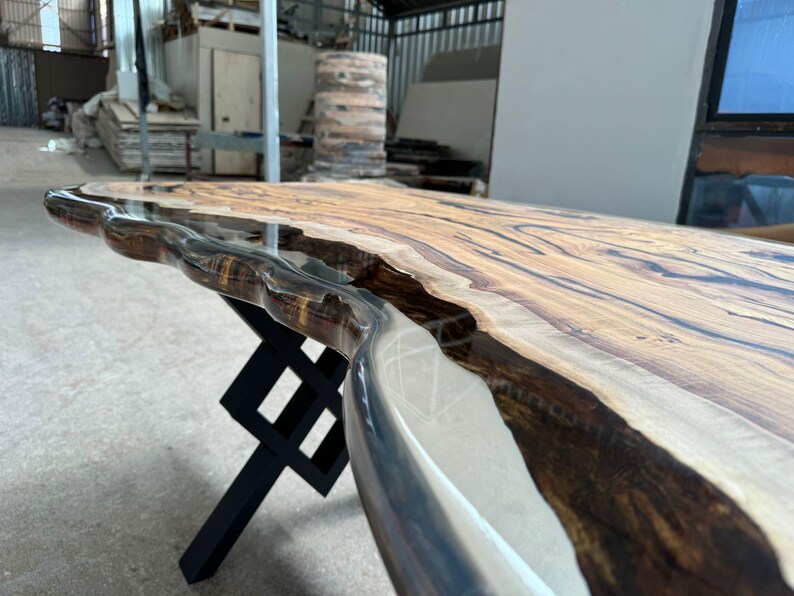 May include: A wooden table top with a live edge and a clear epoxy resin finish. The table top is supported by a black metal X-shaped base.