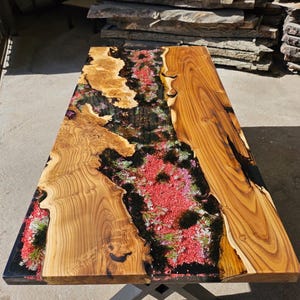 May include: A wooden table top with a natural wood grain finish. The table top has a large resin river with embedded flowers and glitter. The resin is clear and has a high gloss finish.