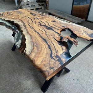 May include: A large wooden table with a live edge top and black epoxy resin filling in the cracks. The table has a clear epoxy resin top and black metal legs.