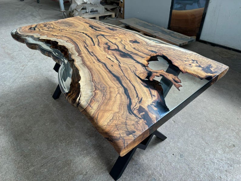May include: A large wooden table with a live edge top and black epoxy resin filling in the cracks. The table has a clear epoxy resin top and black metal legs.