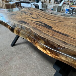 May include: A large, live edge wooden table top with a clear epoxy resin finish. The table top has a natural wood grain pattern and is supported by black metal legs.