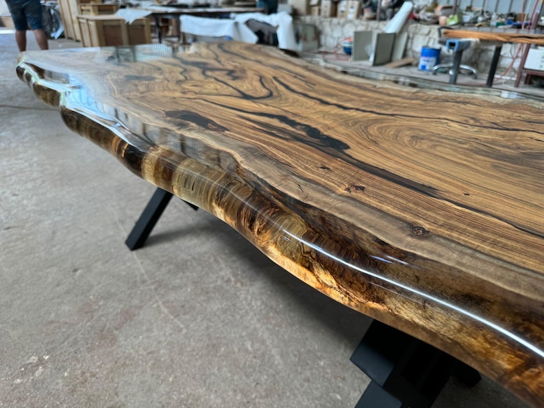 May include: A large, live edge wooden table top with a clear epoxy resin finish. The table top has a natural wood grain pattern and is supported by black metal legs.