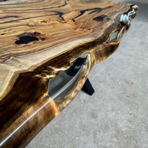 May include: Close-up of a live edge wood table top with a clear epoxy resin fill. The wood has a natural brown and black grain pattern. The epoxy resin is clear and has a slight golden hue.