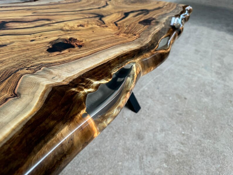 May include: Close-up of a live edge wood table top with a clear epoxy resin fill. The wood has a natural brown and black grain pattern. The epoxy resin is clear and has a slight golden hue.