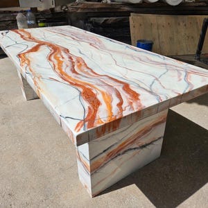 Puede incluir: Una mesa blanca con una tapa de mármol con remolinos naranja y marrón. La mesa tiene una tapa rectangular y dos patas cuadradas.