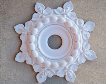 Könnte beinhalten: Weiße Deckenrosette mit einem dekorativen floralen Design und einem zentralen Kreis. Die Rosette hat einen erhabenen, perlenbesetzten Rand und eine glatte, weiße Oberfläche.