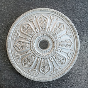 Könnte beinhalten: Eine weiße Deckenrosette mit einem komplizierten floralen Design. Die Rosette hat eine runde Form und eine erhabene Mitte mit einem kleineren Kreis in der Mitte.