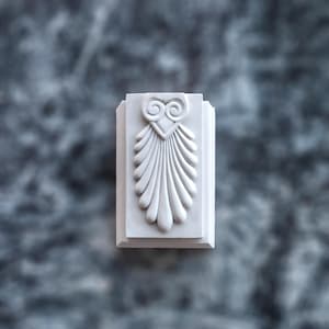 Puede incluir: Un adorno arquitectónico rectangular de yeso blanco que presenta un diseño en relieve central de un motivo estilizado de palmeta u hoja de acanto. La sección superior muestra dos volutas simétricas sobre una serie de formas alargadas.