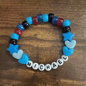 May include: A blue, red and black beaded bracelet with white letter beads spelling out the name "MICHAEL". The bracelet also features blue star and heart charms.