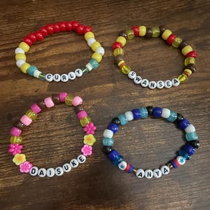 Peut inclure: Quatre bracelets en perles colorées avec différents noms écrits dessus : "Curly", "Swansea", "Daisuke" et "Anya".