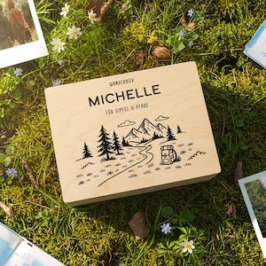 Può includere: Una scatola di legno con il nome "MICHELLE" e un disegno di montagna e sentiero, insieme a due foto polaroid e una mappa, il tutto su un letto di erba verde e piccoli fiori bianchi. La scatola ha il testo "WONDERBOX".