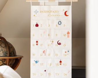 May include: A white fabric Ramadan calendar with gold numbers and crescent moon, star, and mosque icons. The calendar is hanging on a wooden dowel rod. The text "Ramadan Kareem" is printed in gold at the top.