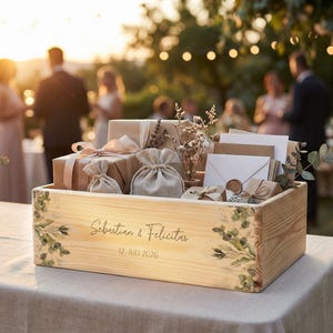 Könnte beinhalten: Holzkiste gefüllt mit Geschenken, Umschlägen und kleinen Beuteln, verziert mit floralen Akzenten. Die Kiste trägt die Namen "Sebastian & Felicitas" und das Datum "12. Juli 2026". Die Szene spielt sich im Freien bei einer Hochzeit ab.