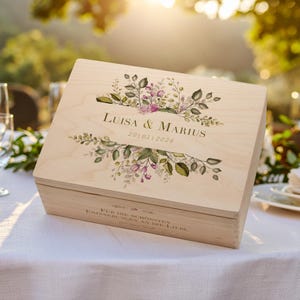 Puede incluir: Una caja de madera clara con un diseño floral y los nombres "Luisa & Marius" y la fecha "201012026" impresos en la tapa. La caja es rectangular y está sobre un mantel blanco.