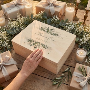 AMARI Erinnerungskiste Hochzeit Pflanzenwelt von oben: eine Hand liegt auf dem bedruckten Deckel, daneben verpackte Geschenke und Kerzen.