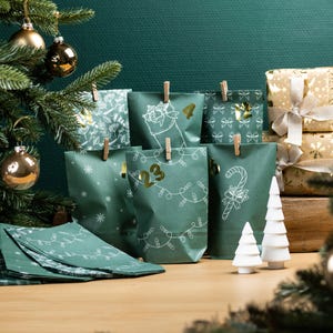 Peut inclure: Calendrier de l'Avent avec des sacs en papier verts décorés d'illustrations blanches et de chiffres dorés. Les sacs sont fixés avec des pinces à linge en bois. Des sapins de Noël en céramique blanche et des cadeaux emballés sont également visibles.