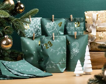 May include: An advent calendar with green paper bags decorated with white illustrations and gold numbers. The bags are clipped with wooden clothespins. White ceramic Christmas trees and wrapped gifts are also visible.