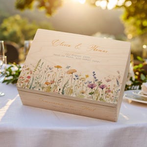 Può includere: Una scatola di legno con un design floreale e i nomi "Olivia & Thomas" in oro. La scatola ha la data "12 agosto 2025" e la frase "Hand in Hand, ein Leben lang." La scatola è su una tovaglia bianca.